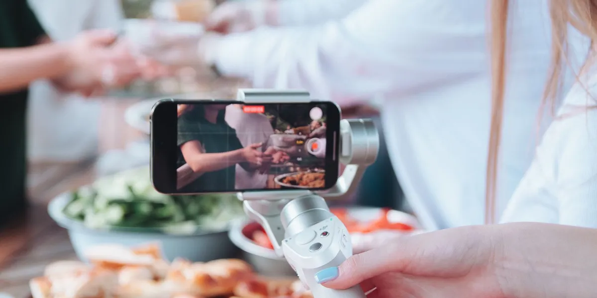 A woman's hand holds a smartphone on a white handheld gimbal, recording people serving food at an outdoor table; the phone screen shows the scene with a red recording timer.