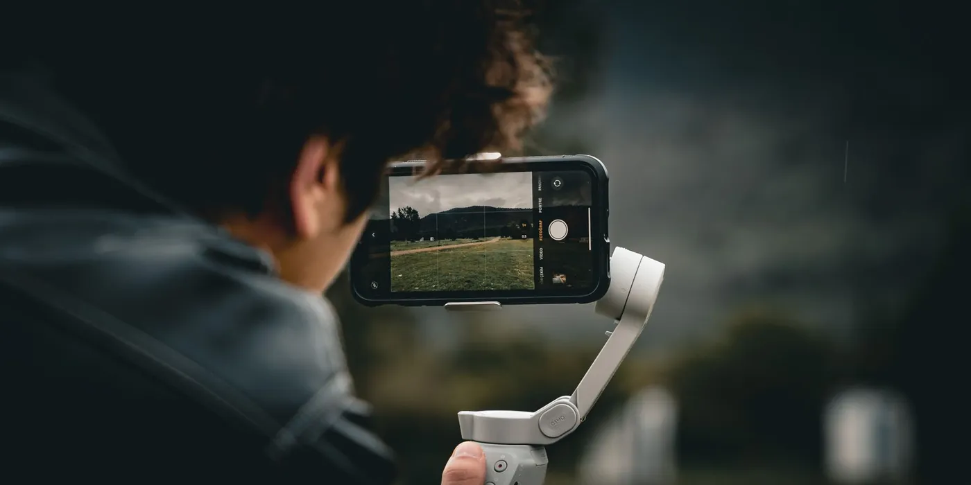 Seen from over his shoulder, a man holds a smartphone on a handheld gimbal, its screen framing a grassy field and hills under a cloudy sky.
